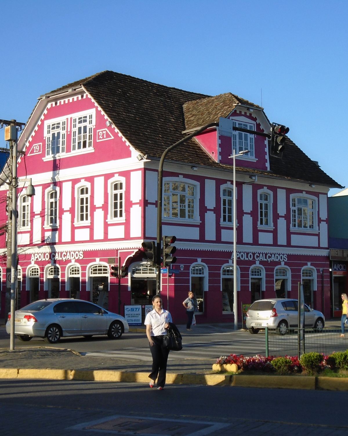 Pedestres atravessando a rua diante de um casarão pintado de rosa e branco, com telhado alto e janelas enfileiradas, no Centro de Joinville, no fim da tarde de sol.