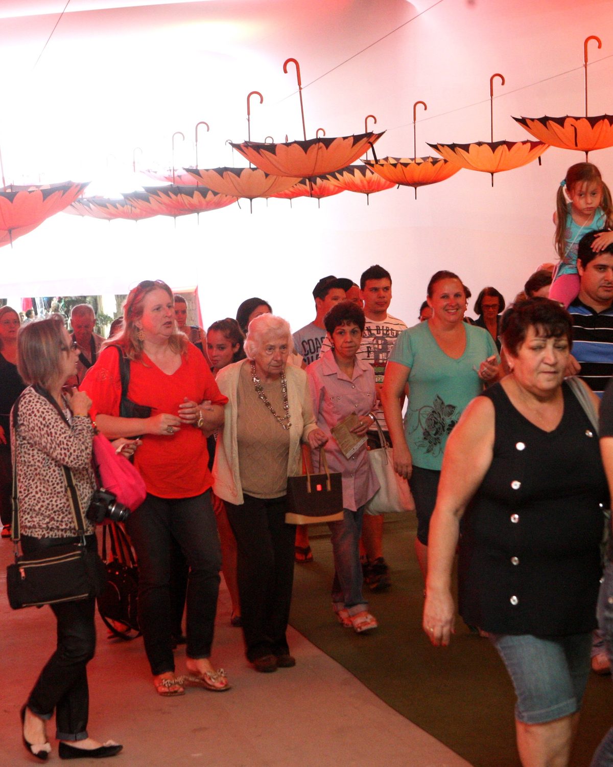 Visitantes adultos caminhando por um corredor iluminado de vermelho, com guarda-chuvas abertos pendurados no teto, na Festa das Flores, na Expoville, em Joinville.