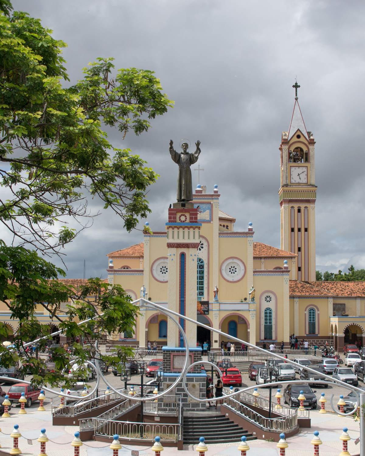 Santuário de São Francisco das Chagas, em Juazeiro do Norte, com a fachada amarela, a torre do relógio e a estátua de São Francisco de braços abertos sobre a praça cheia de carros estacionados, sob céu nublado.