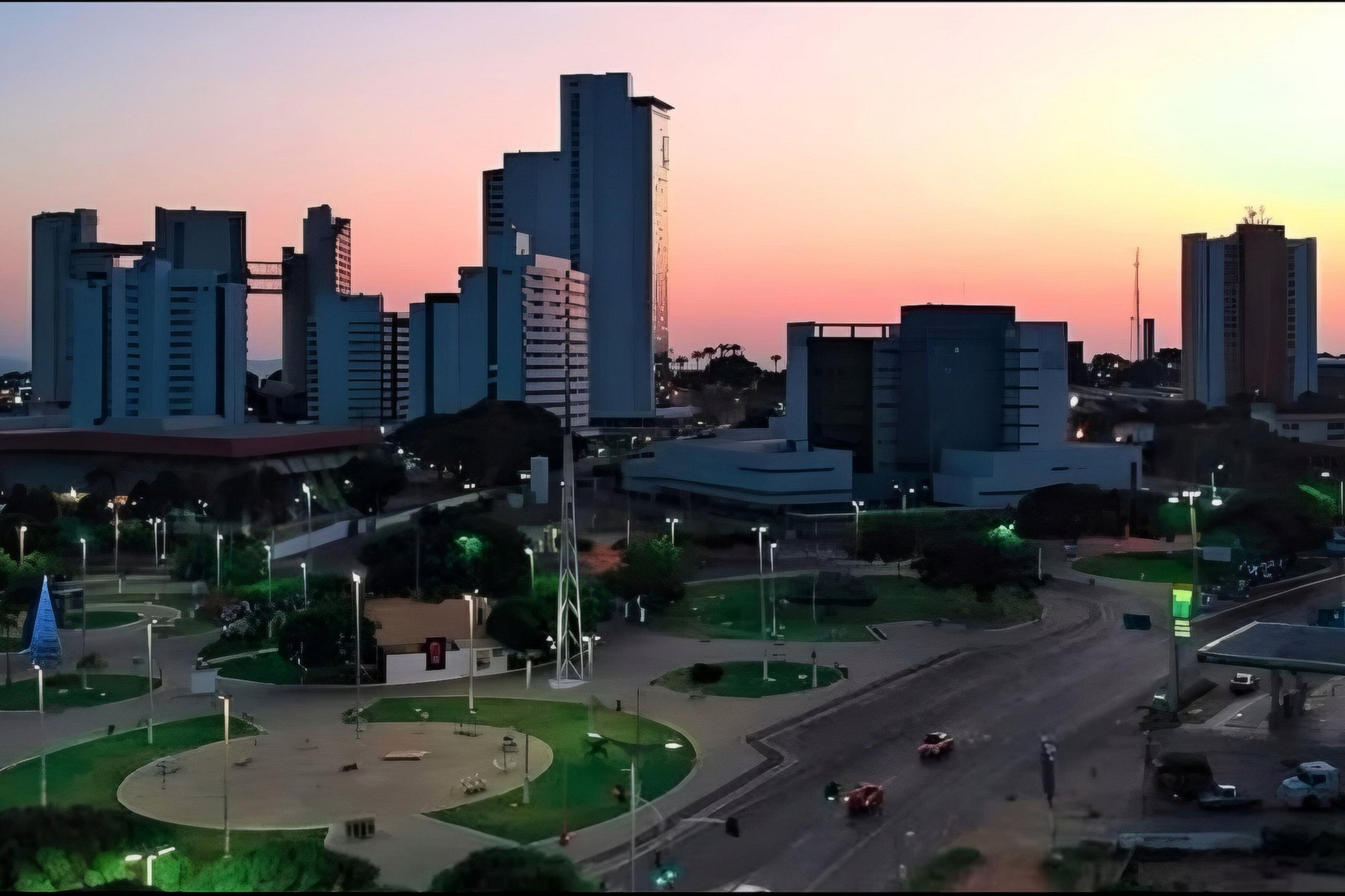 Anoitecer na Praça do Giradouro, no bairro Triângulo, em Juazeiro do Norte, com os canteiros e postes acesos, a avenida com carros e os prédios altos recortados contra o céu rosa e laranja.