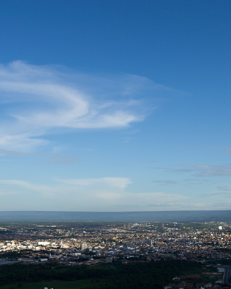 Vista de Juazeiro do Norte do alto da Colina do Horto, com a cidade espalhada pela planície até a linha da serra no horizonte, sob céu azul com nuvens finas.