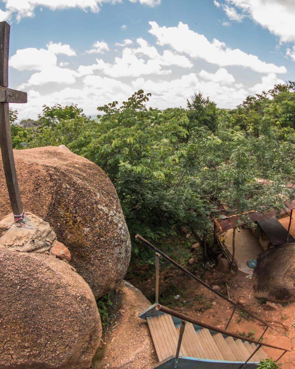 Cruz de madeira fincada sobre grandes blocos de granito arredondados no Santo Sepulcro, em Juazeiro do Norte, com a mata verde em volta, a escada da trilha lá embaixo e o céu azul com nuvens brancas.