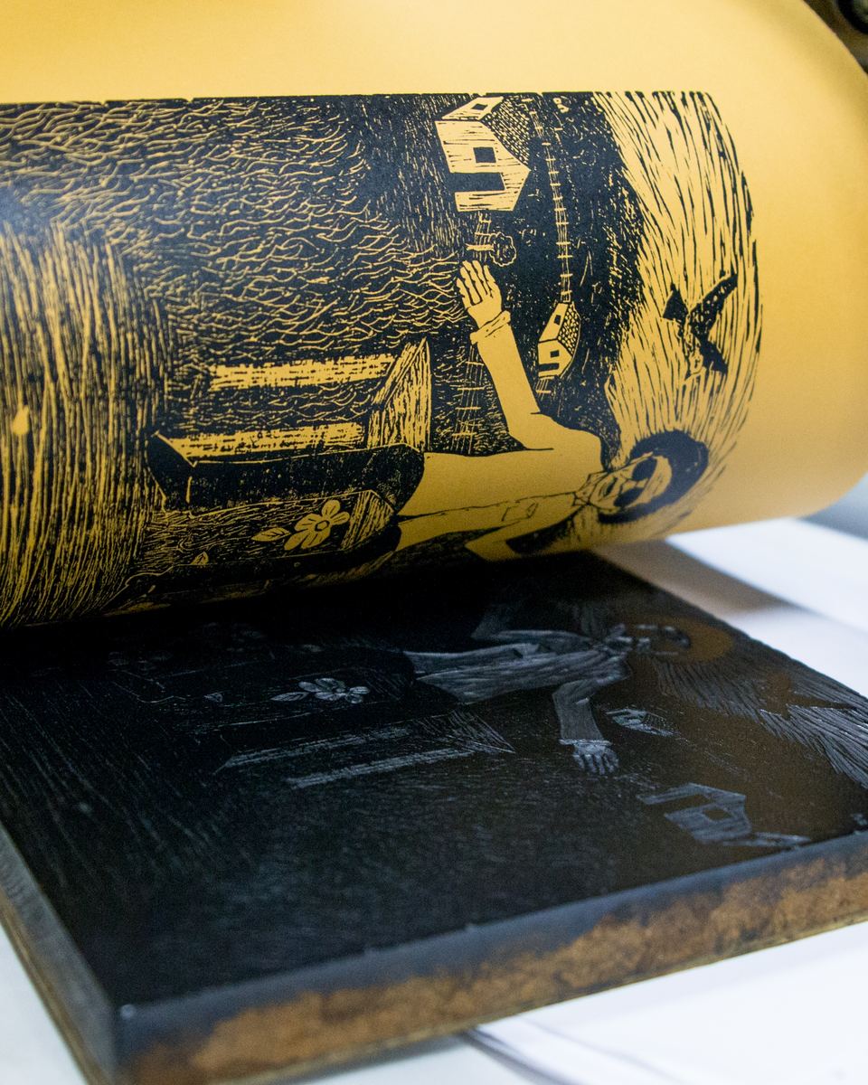 Folha de papel amarelo sendo levantada de uma matriz de xilogravura entintada de preto, revelando a gravura de uma figura e um pássaro, numa prensa da Lira Nordestina.