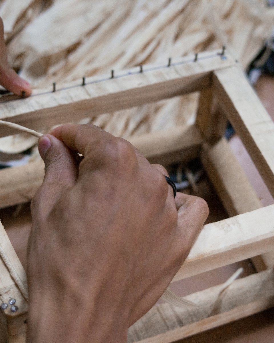 Mãos passando uma fibra por uma pequena armação de madeira com pregos, ao lado de uma vasilha azul com água e de fibras claras, na Associação dos Artesãos da Mãe das Dores.