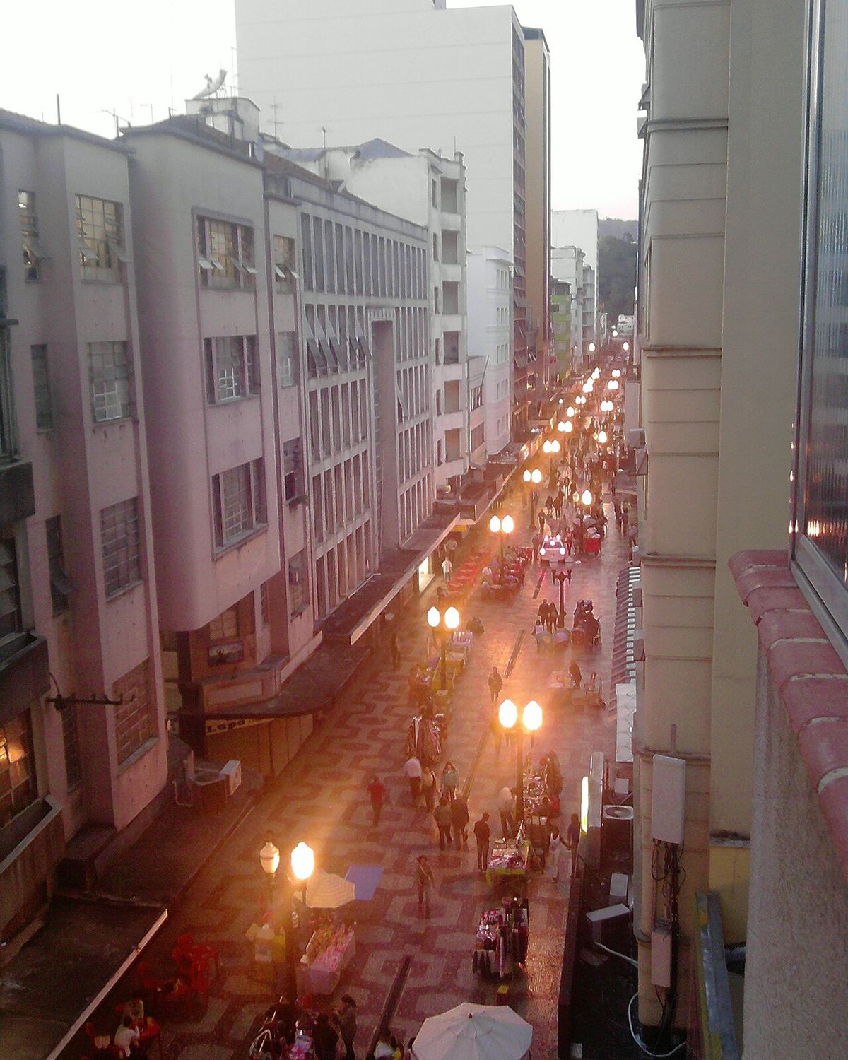 Calçadão da Rua Halfeld visto do alto no fim da tarde, cheio de pedestres entre barracas e postes acesos, com prédios dos dois lados, no centro de Juiz de Fora.