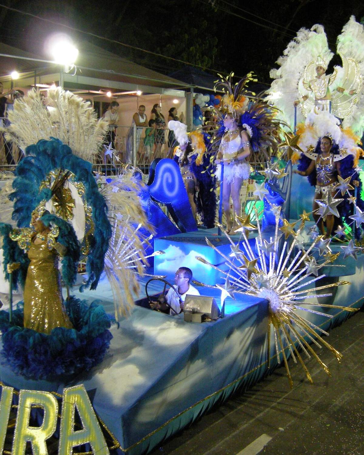 Carro alegórico azul e prateado com destaques de fantasia de plumas desfilando à noite no carnaval de Juiz de Fora, com o público na arquibancada e a mão de um espectador segurando o celular em primeiro plano.