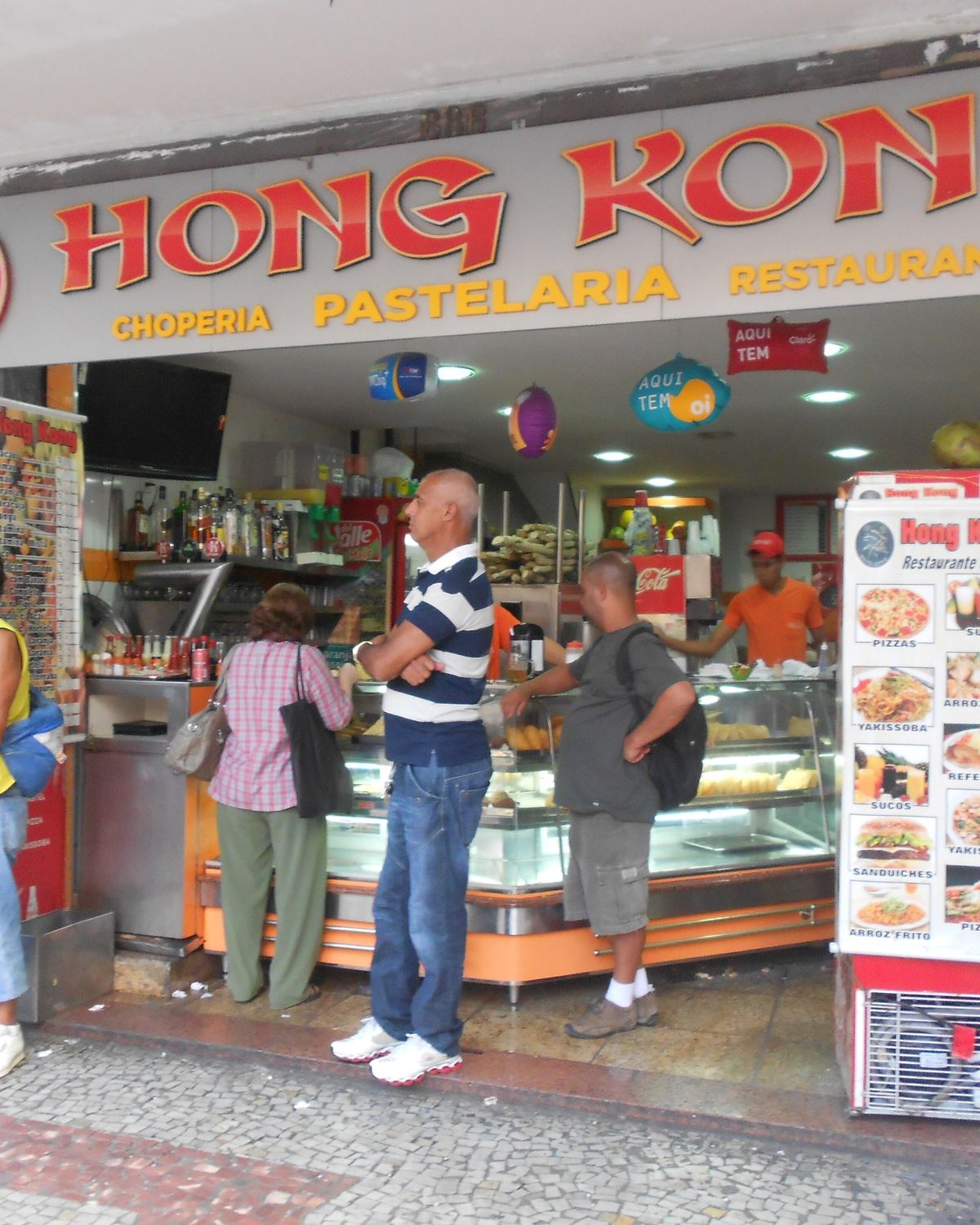 Clientes em pé no balcão de uma pastelaria e choperia do centro de Juiz de Fora, um deles com um copo de chope na mão, diante da vitrine de salgados.