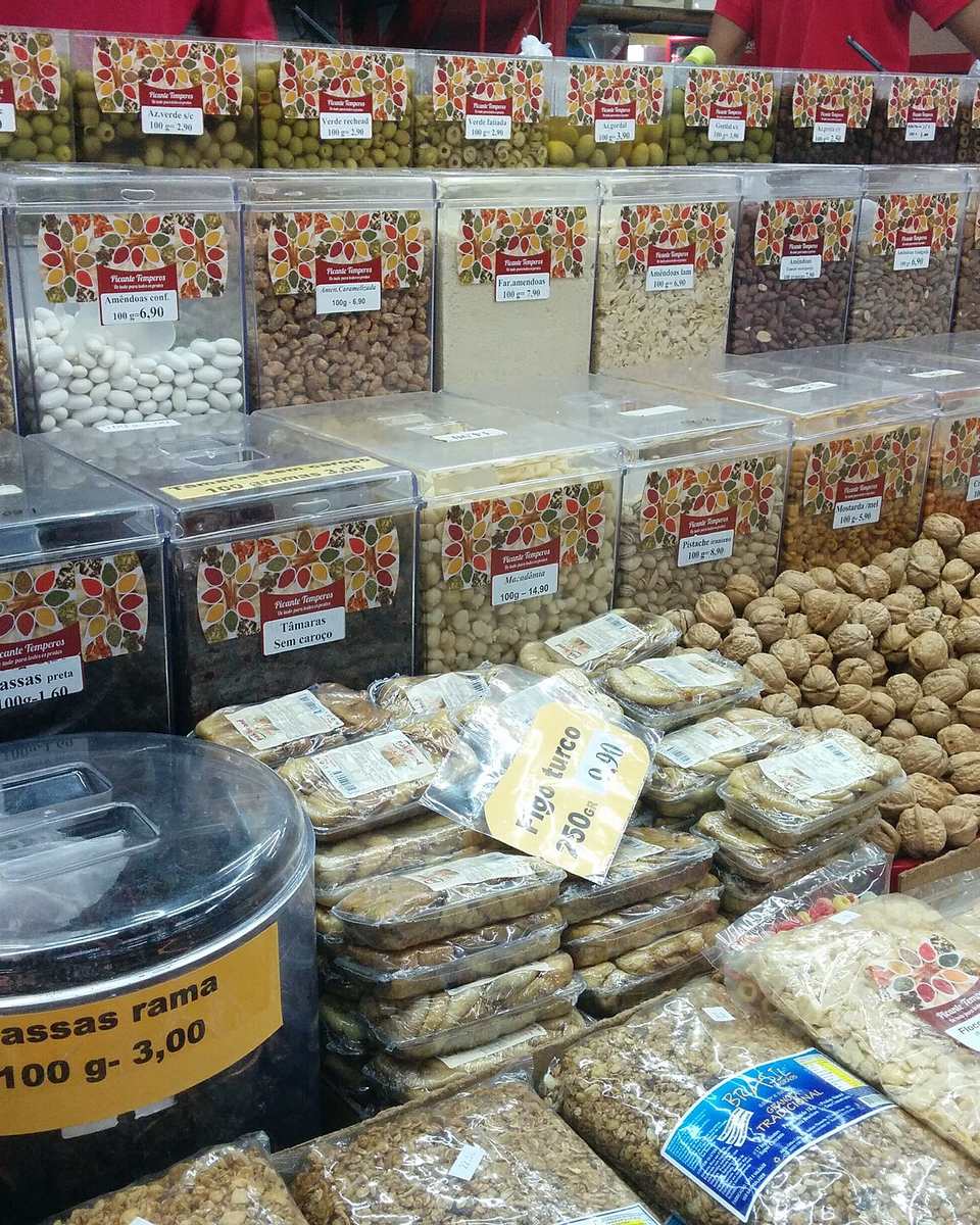 Banca do Mercado Municipal de Juiz de Fora com potes de castanhas, nozes, tâmaras, uvas-passas e azeitonas vendidos a granel, com preços em plaquinhas e atendentes de camiseta vermelha ao fundo.