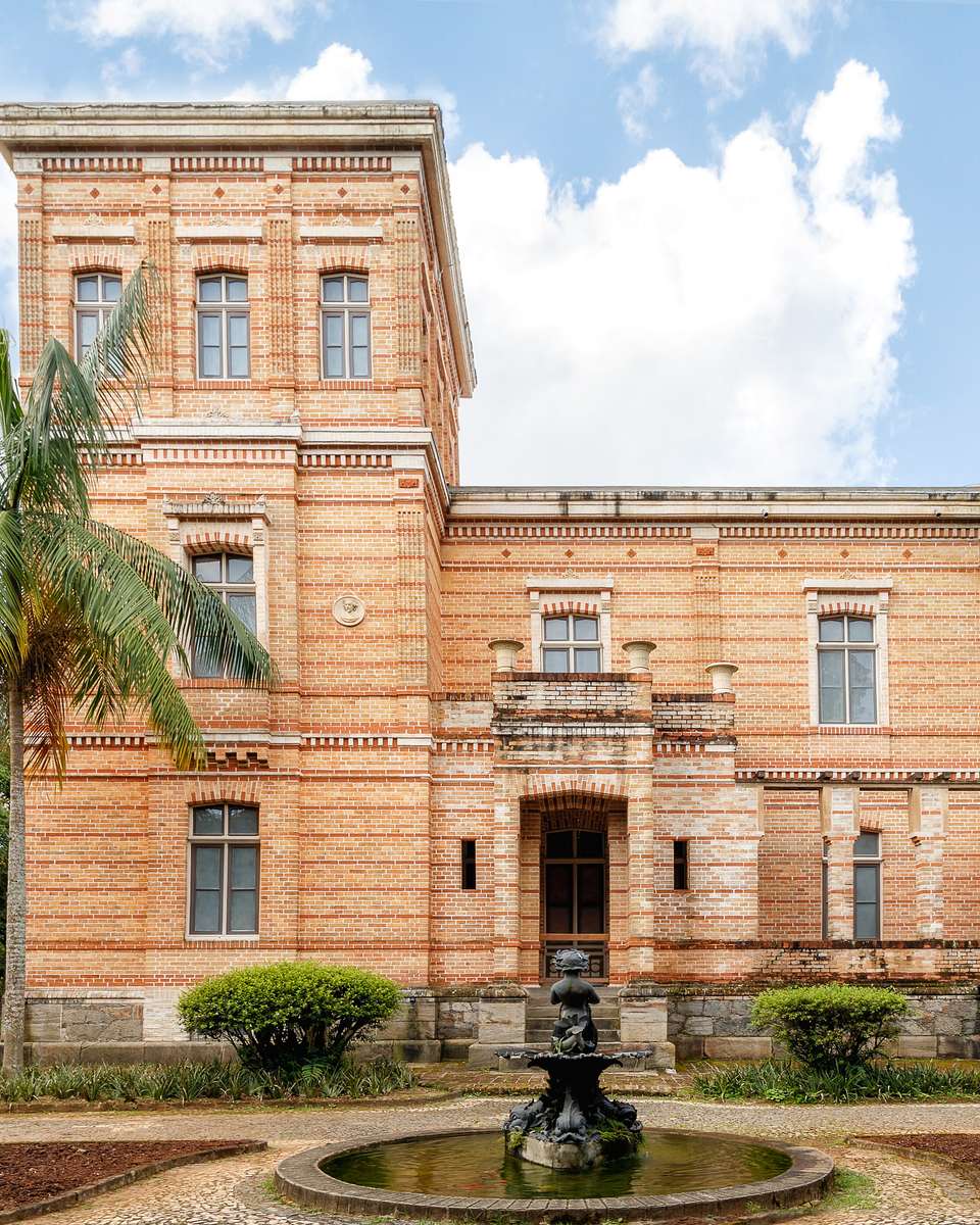 Prédio de tijolos aparentes do Museu Mariano Procópio, em Juiz de Fora, com palmeira, jardim e chafariz na frente, sob céu azul com nuvens.