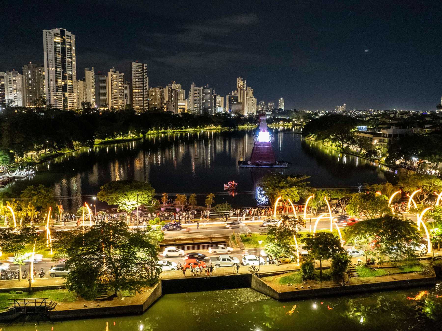 Lago Igapó à noite, com as árvores da margem enfeitadas de luzes de Natal refletidas na água e os prédios iluminados ao fundo, em Londrina.