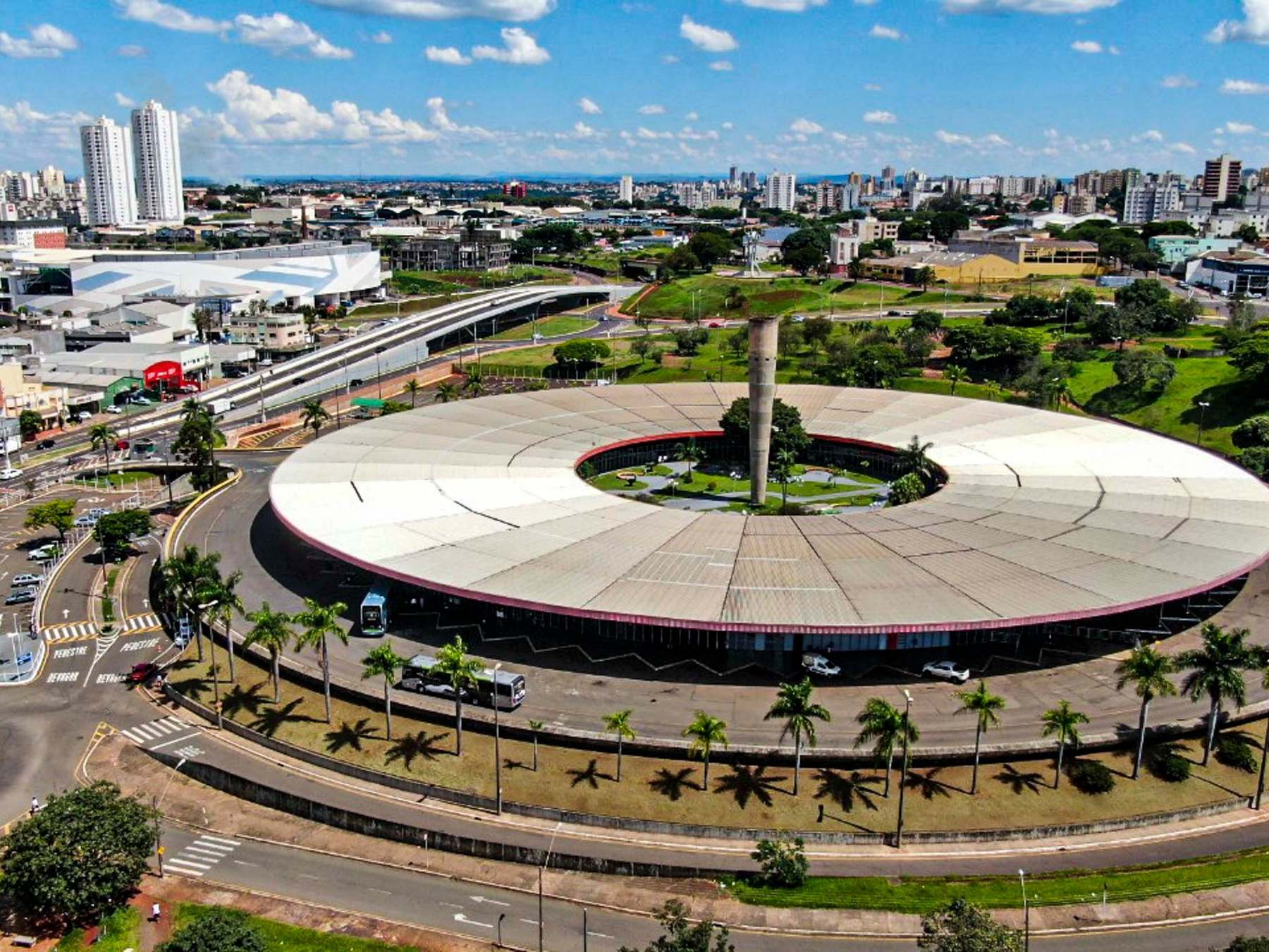 Vista aérea do Terminal Rodoviário de Londrina, um grande prédio circular de cobertura clara com um jardim no centro, cercado por palmeiras e avenidas, sob céu azul com nuvens.