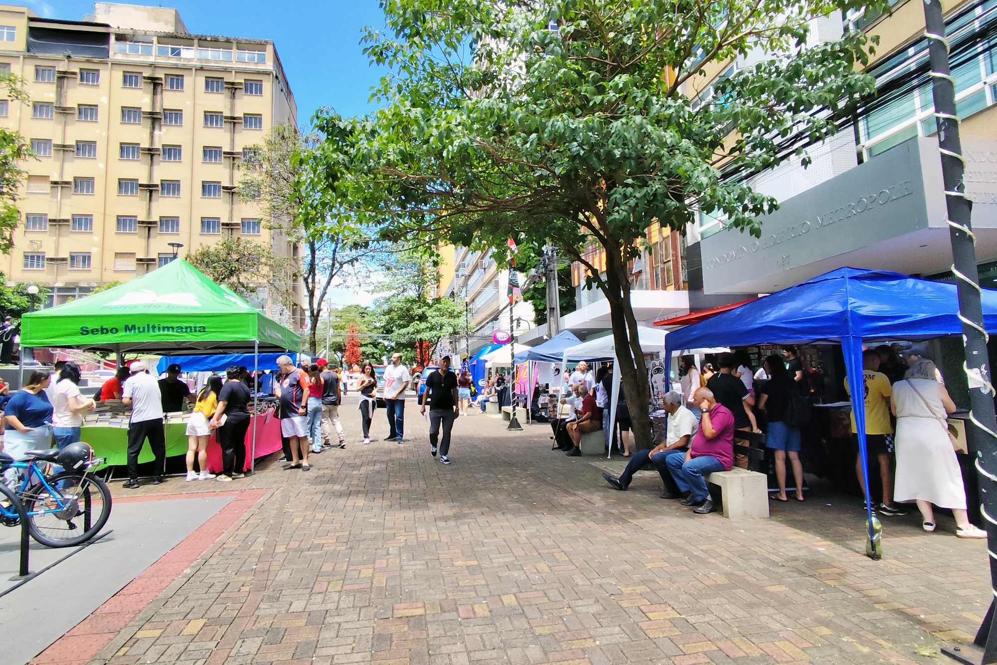 Pessoas caminhando entre as barracas de uma feira de livros no Calçadão de Londrina, com tendas verdes e azuis, árvores e prédios do Centro num dia de sol.