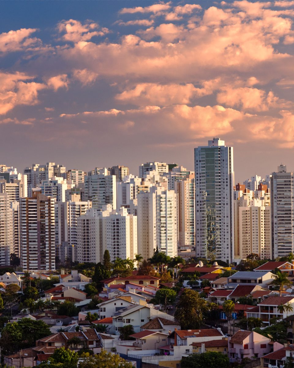 Prédios altos de Londrina iluminados pelo sol do fim de tarde, com casas de telhado vermelho e árvores na frente e nuvens rosadas no céu.