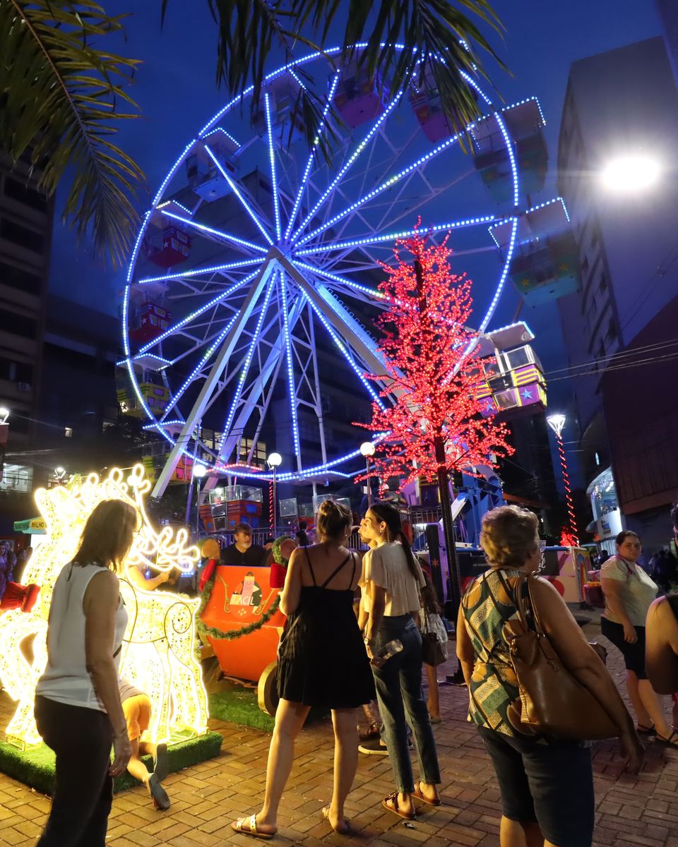 Roda-gigante iluminada de azul e uma árvore de luzes vermelhas no Calçadão de Londrina à noite, com moradores passeando e olhando a decoração de Natal.