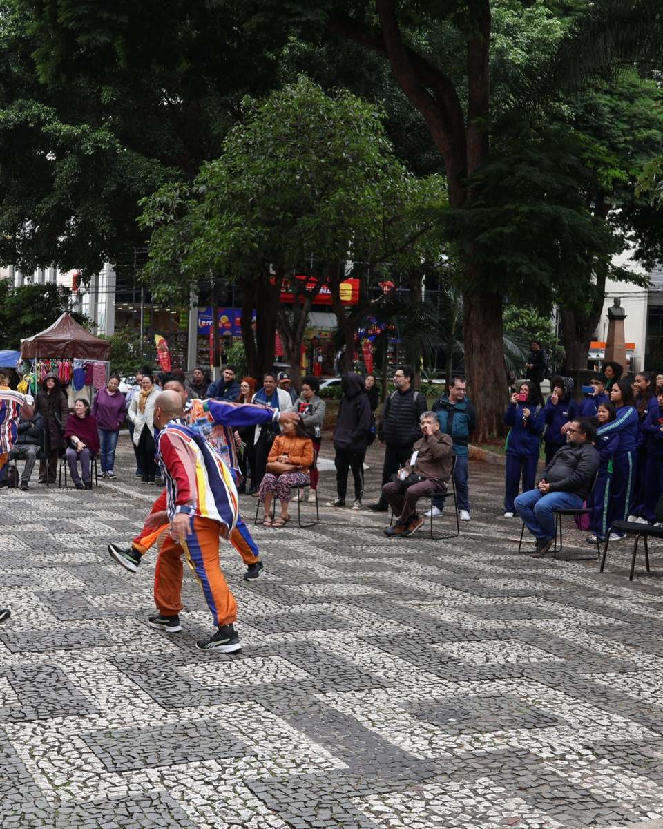Artistas com roupas listradas coloridas se apresentando no piso de pedra portuguesa do Calçadão de Londrina durante o FILO, com o público sentado em cadeiras e em pé sob as árvores.