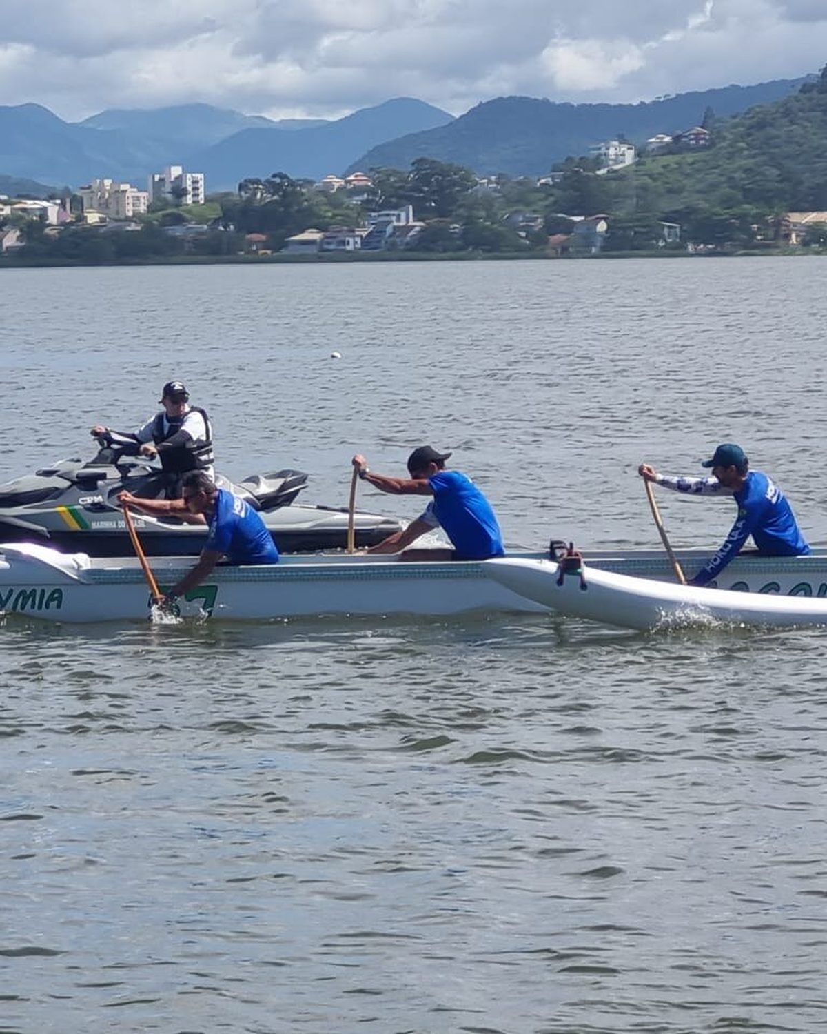 Quatro remadores de camiseta azul numa canoa havaiana na Lagoa de Imboassica, em Macaé, acompanhados por uma moto aquática da Marinha, com casas, prédios e montanhas ao fundo.