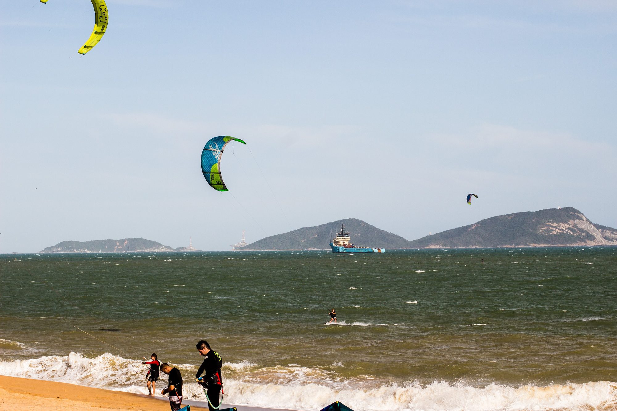 Kitesurfistas na beira da praia em Macaé, com um barco de apoio offshore e plataformas no horizonte.