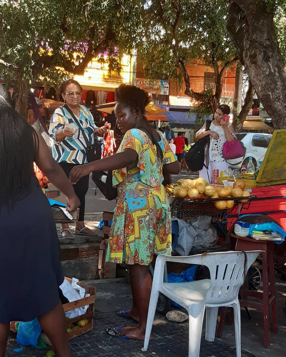 Feira de rua em Manaus, com vendedoras atendendo na banca de frutas embaixo das árvores e uma cliente conferindo o celular.