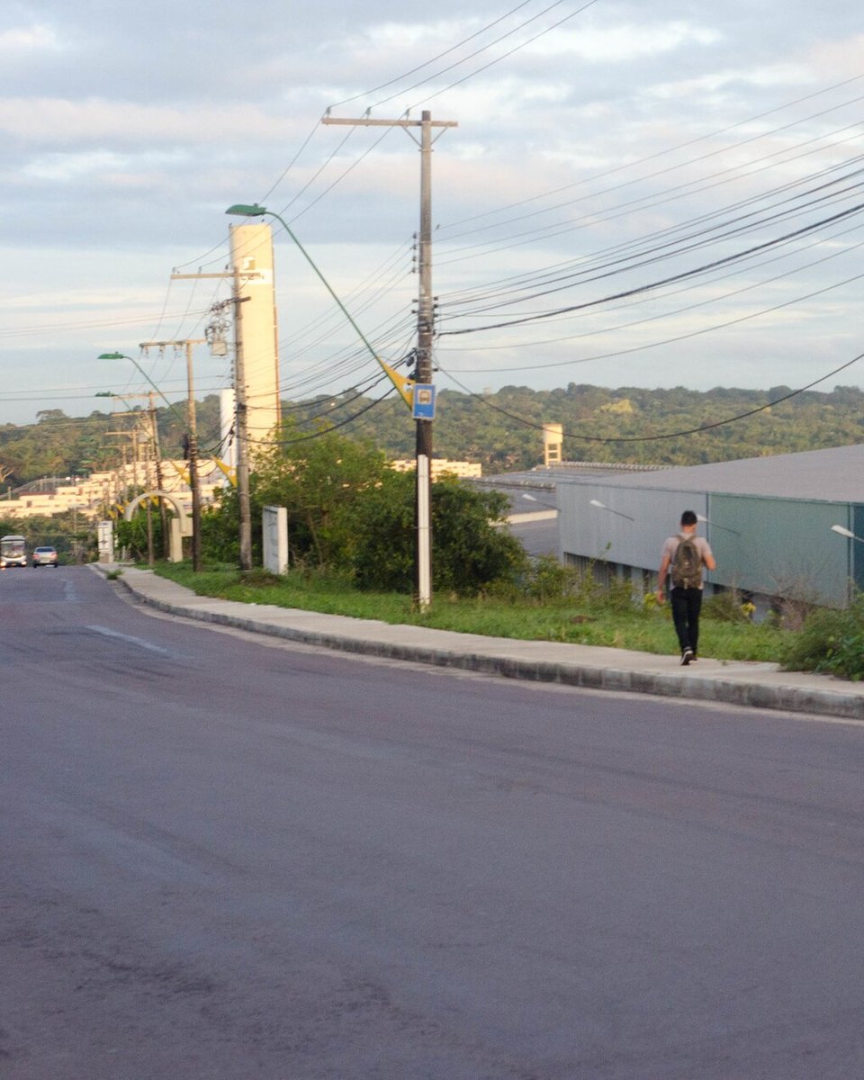 Avenida do Distrito Industrial de Manaus no fim da tarde, com galpões de fábrica ao lado da pista e um trabalhador caminhando pela calçada.