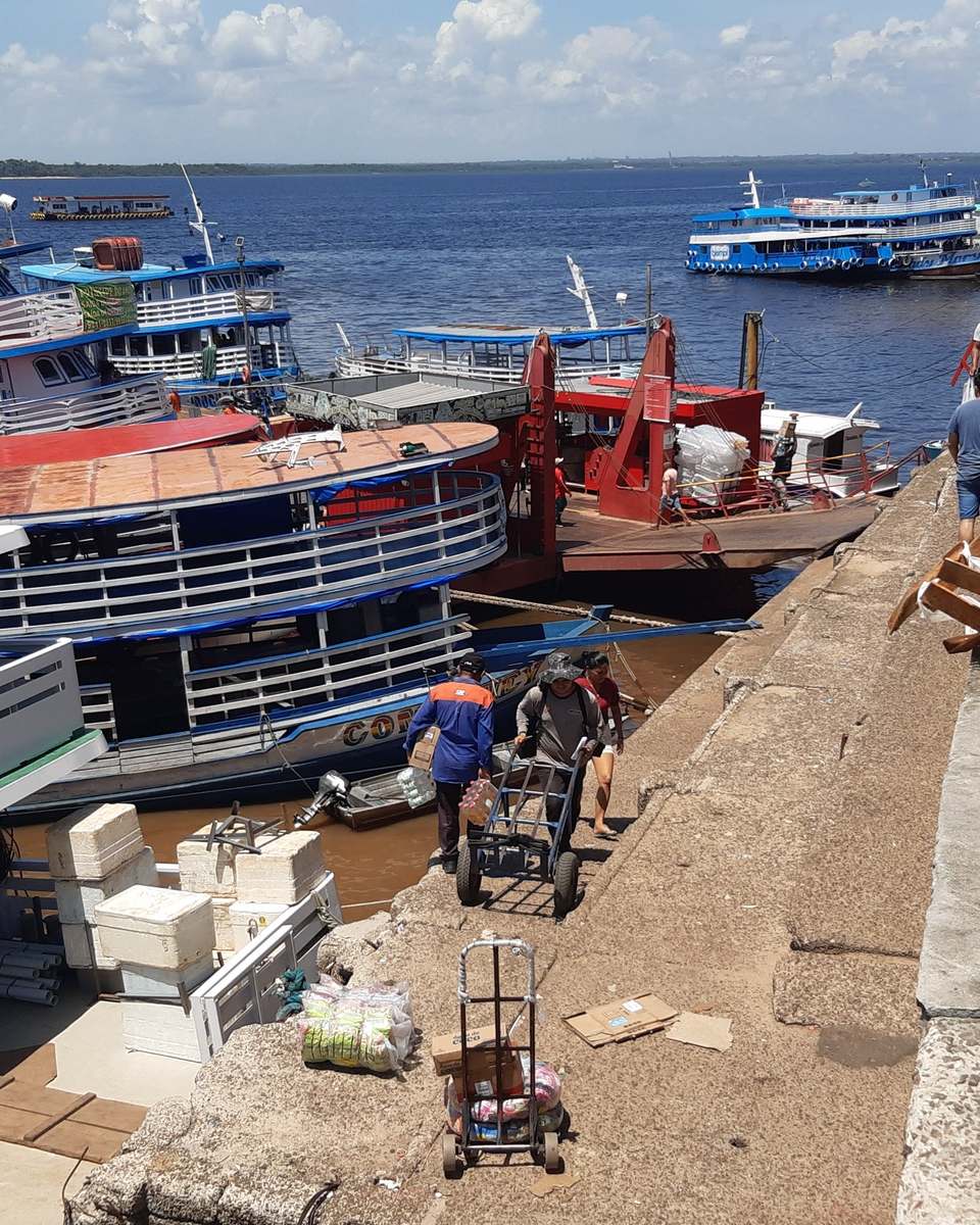 Barcos regionais de carga atracados na orla do centro de Manaus, com carregadores subindo a rampa com carrinhos de mão e motos estacionadas na beira.
