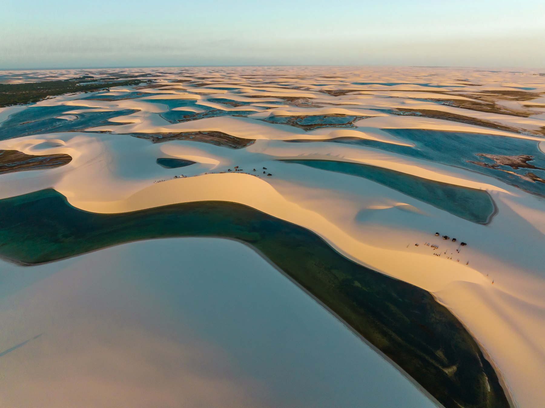Mar de dunas dos Lençóis Maranhenses visto do alto em Santo Amaro do Maranhão, com faixas de areia clara separando lagoas de água escura até o horizonte, na luz do fim de tarde