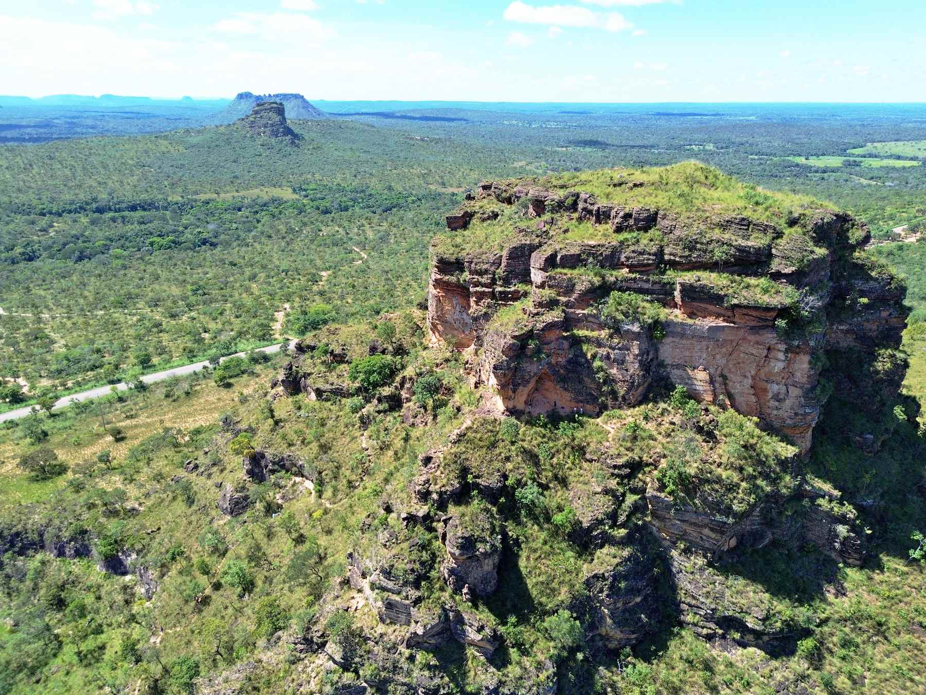 Formação de arenito em forma de mesa na Chapada das Mesas vista do alto, com o paredão avermelhado coberto de vegetação verde, o cerrado em volta e outro morro chato ao longe