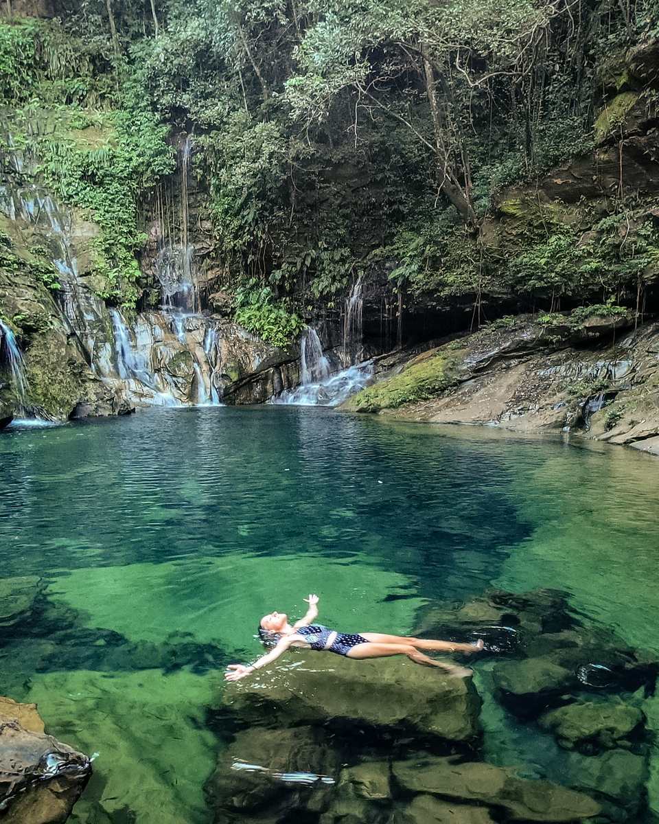 Poço de água cristalina verde e azul cercado por paredão de rocha e mata, com pequenas quedas d'água ao fundo e uma pessoa boiando