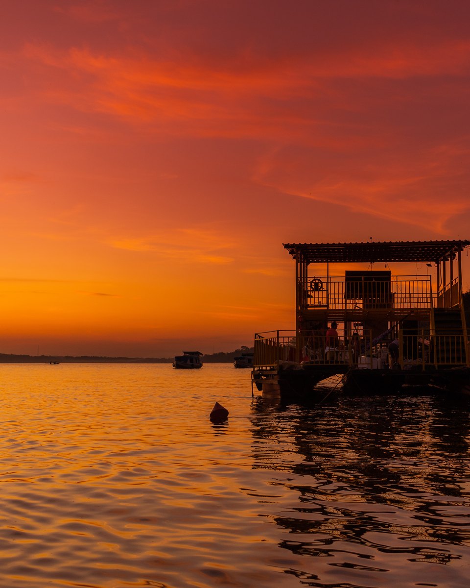 Fim de tarde no rio Tocantins em Imperatriz, com um bar flutuante de madeira ancorado na margem, barcos na água e os prédios da cidade recortados contra o céu alaranjado