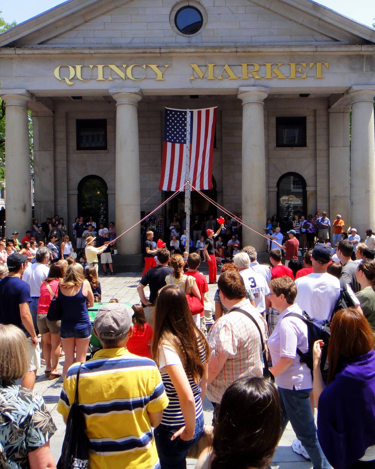 Multidão reunida em frente ao Quincy Market, em Boston, assistindo a uma apresentação de rua num dia de sol, com a bandeira americana entre as colunas.