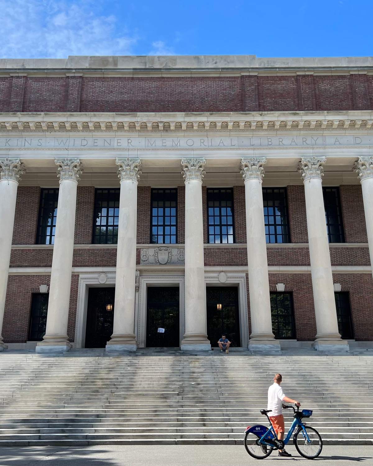 Homem com uma bicicleta compartilhada parado diante da escadaria e das colunas da Biblioteca Widener, na Universidade Harvard, em Cambridge.