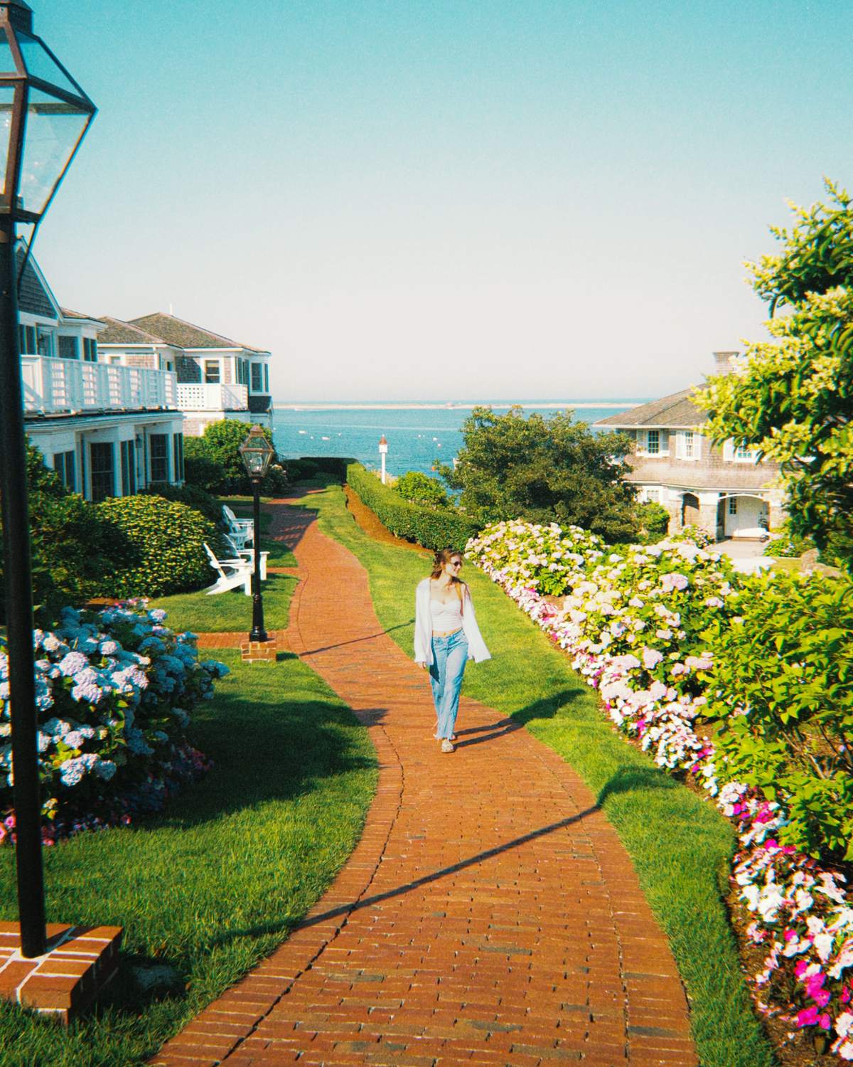 Mulher de camisa branca e jeans caminhando por um caminho de tijolos entre hortênsias floridas, com casas de madeira e o mar ao fundo, em Chatham, no Cape Cod.