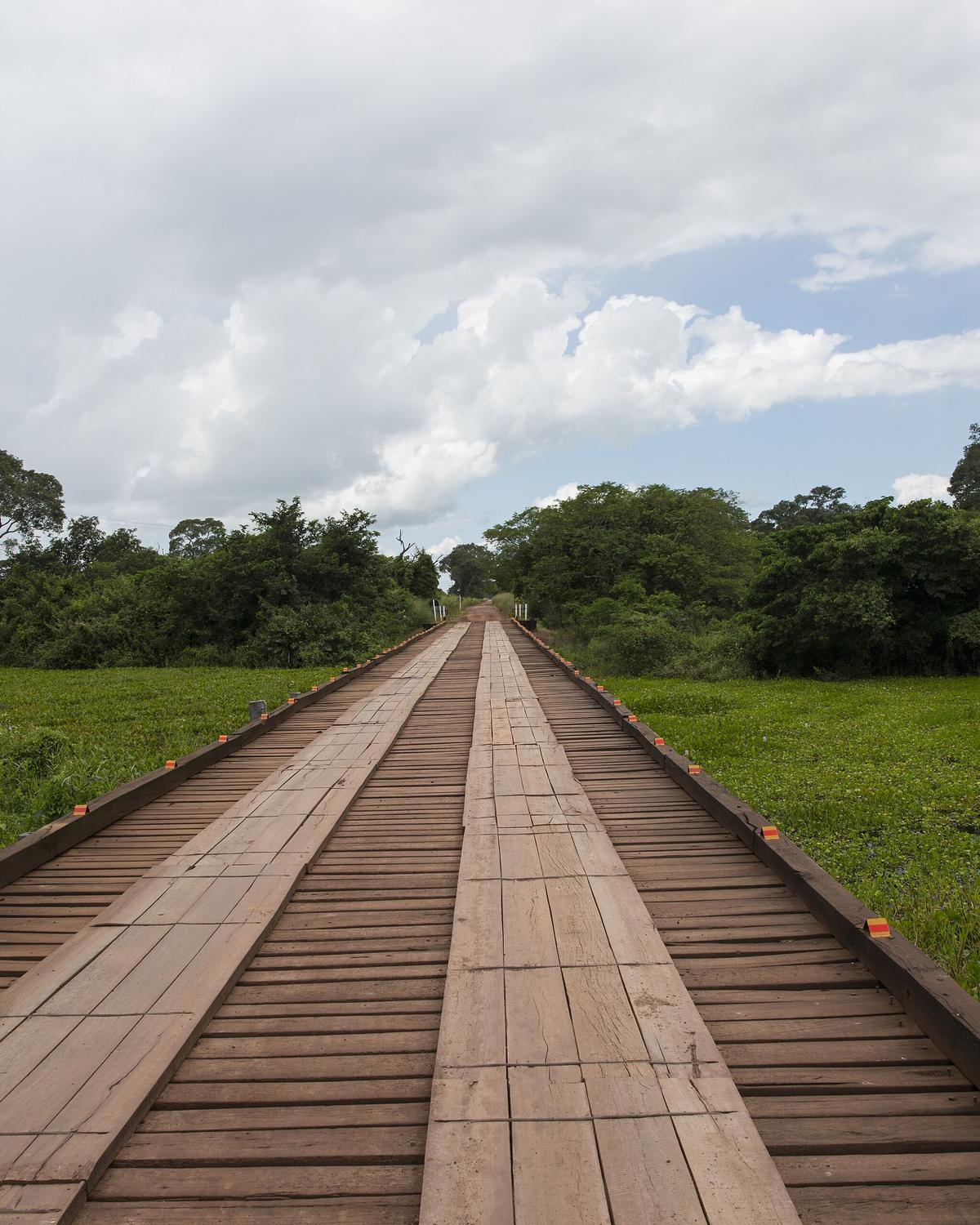 Ponte de madeira da Estrada Parque do Pantanal seguindo reta sobre a planície verde alagada, com céu carregado de nuvens.