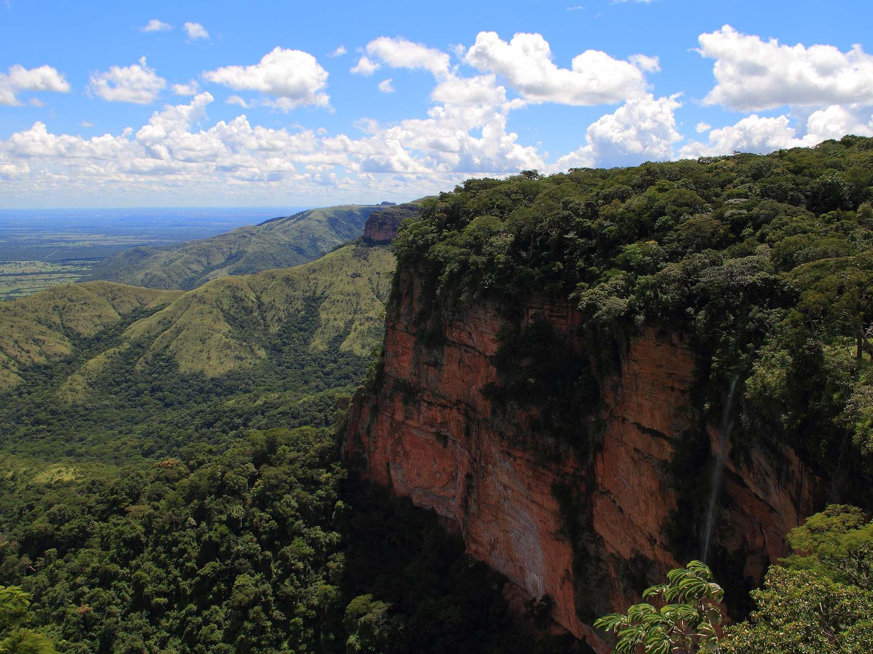 Vista do Mirante do Morro dos Ventos na Chapada dos Guimarães, com o paredão de rocha vermelha coberto de mata, o vale verde à esquerda e o céu azul com nuvens brancas