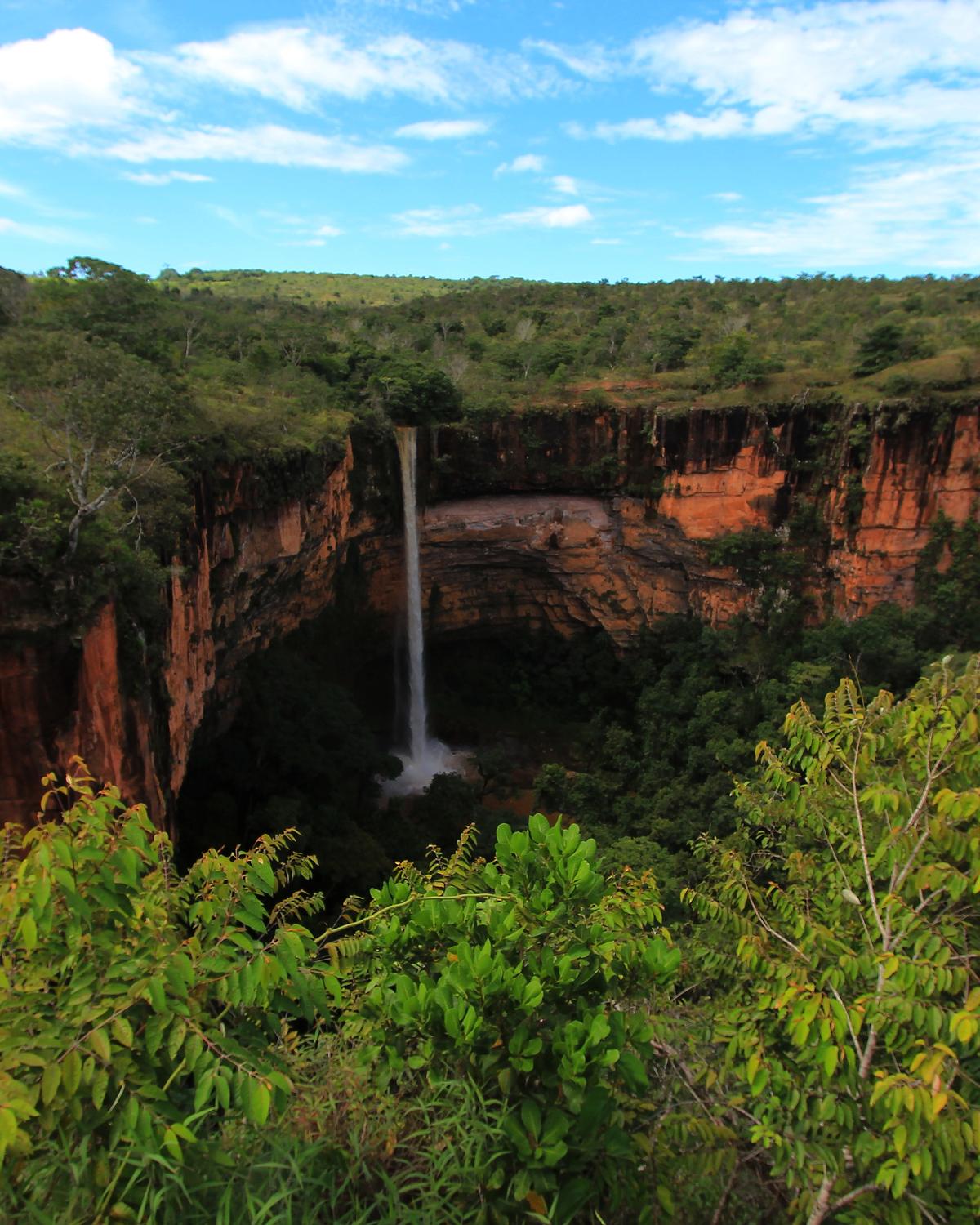 Cachoeira Véu de Noiva caindo do paredão vermelho do anfiteatro rochoso da Chapada dos Guimarães, cercada de vegetação verde, sob céu azul.