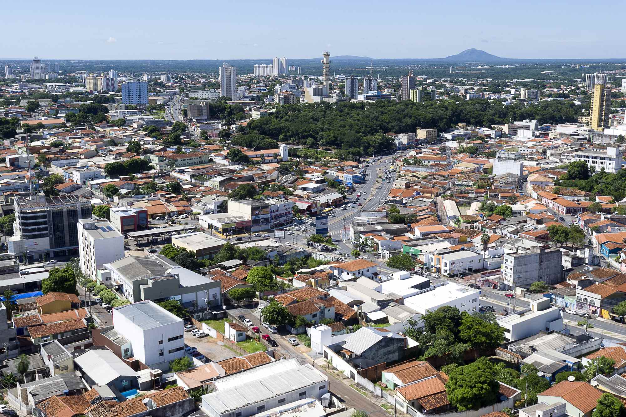 Vista aérea do centro de Cuiabá em dia de céu azul, com prédios altos, casas de telhado vermelho e o Morro de Santo Antônio ao fundo.