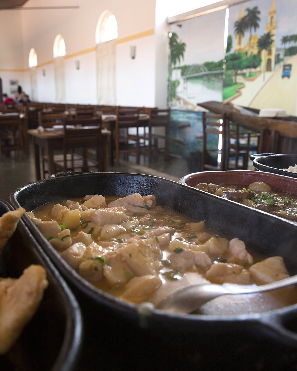 Travessas de ferro com peixe frito e peixe ensopado servidas em um restaurante de comida regional em Cuiabá, com mesas, clientes e um painel pintado da cidade ao fundo.