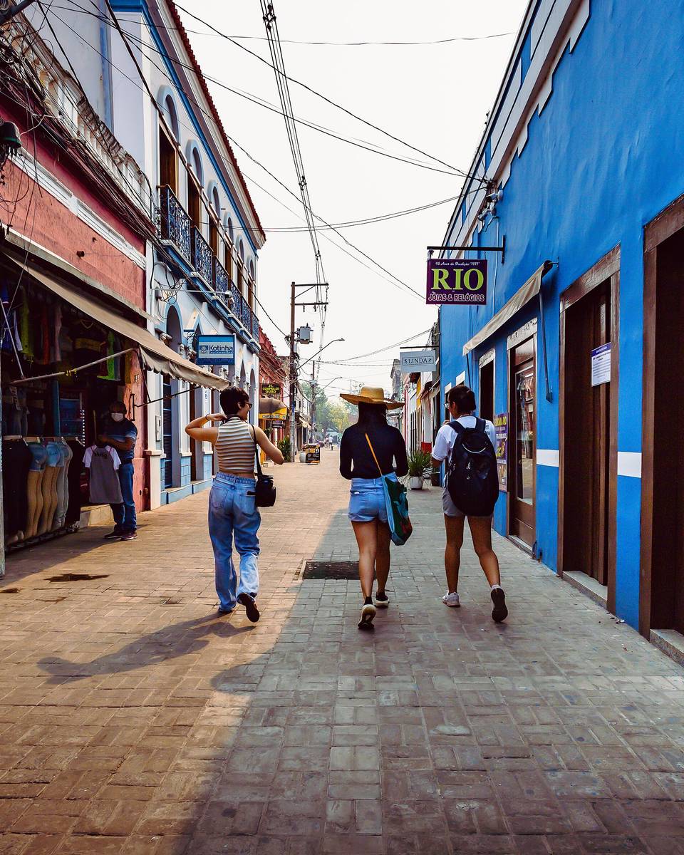 Três pessoas caminhando por uma rua estreita de comércio no centro histórico de Cuiabá, entre casarões pintados de rosa e azul com portas de madeira e lojas abertas.