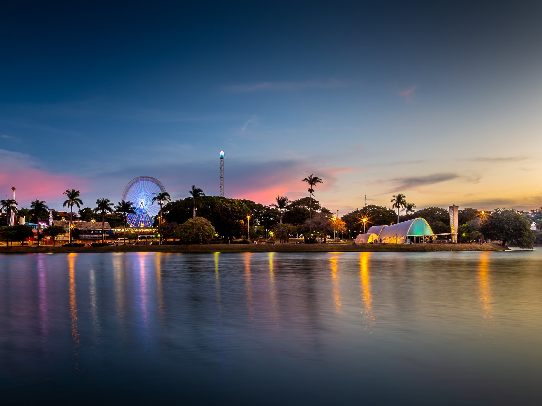 Lagoa da Pampulha ao entardecer, com as luzes da orla refletidas na água, a roda-gigante e a Igreja de São Francisco de Assis iluminada.