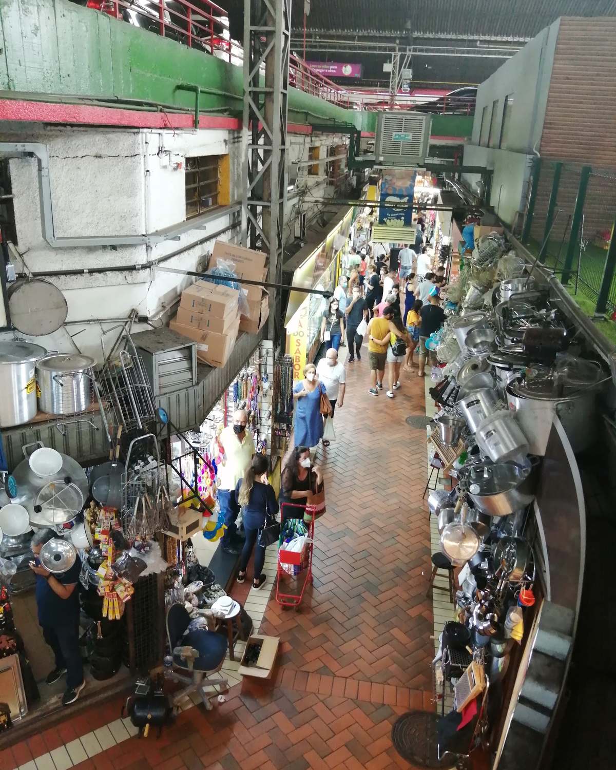 Corredor do Mercado Central de Belo Horizonte visto do alto, cheio de gente entre bancas de panelas, utensílios e produtos mineiros.