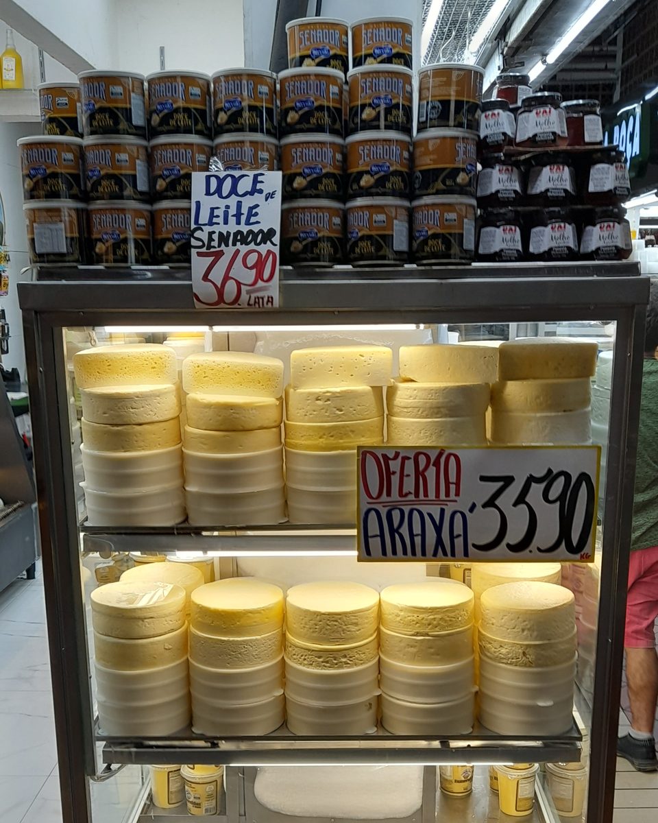 Vitrine refrigerada com queijos Minas artesanais empilhados e placa de oferta do queijo de Araxá, com latas de doce de leite em cima, no Mercado Central de Belo Horizonte.