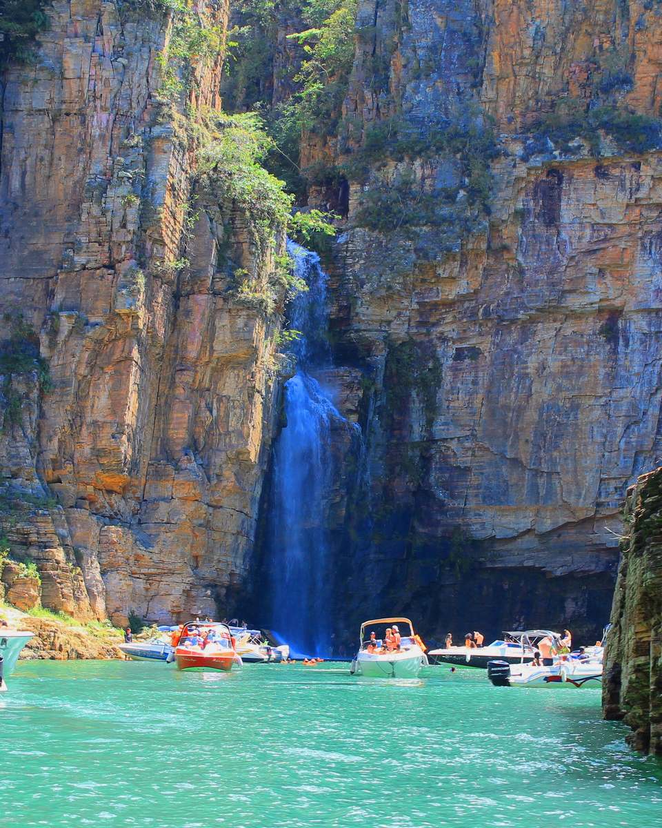 Lanchas cheias de turistas na água verde do Lago de Furnas, diante de uma cachoeira que desce pelo paredão dos cânions de Capitólio.
