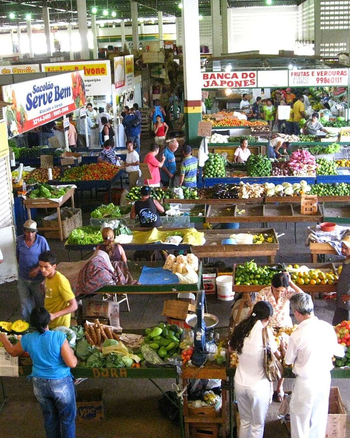 Corredores do Mercado Municipal de Montes Claros vistos do alto, com bancas de frutas, verduras e legumes, clientes escolhendo produtos e comerciantes atrás dos balcões.