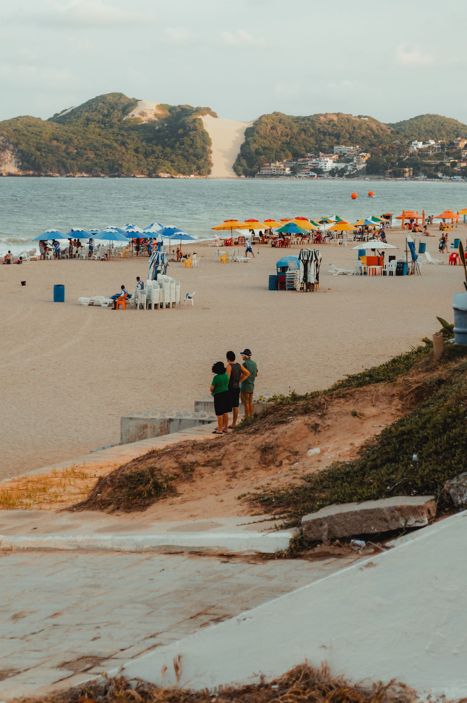 Faixa de areia da praia de Ponta Negra com guarda-sóis coloridos e cadeiras, moradores conversando na beira da duna, com o Morro do Careca ao fundo