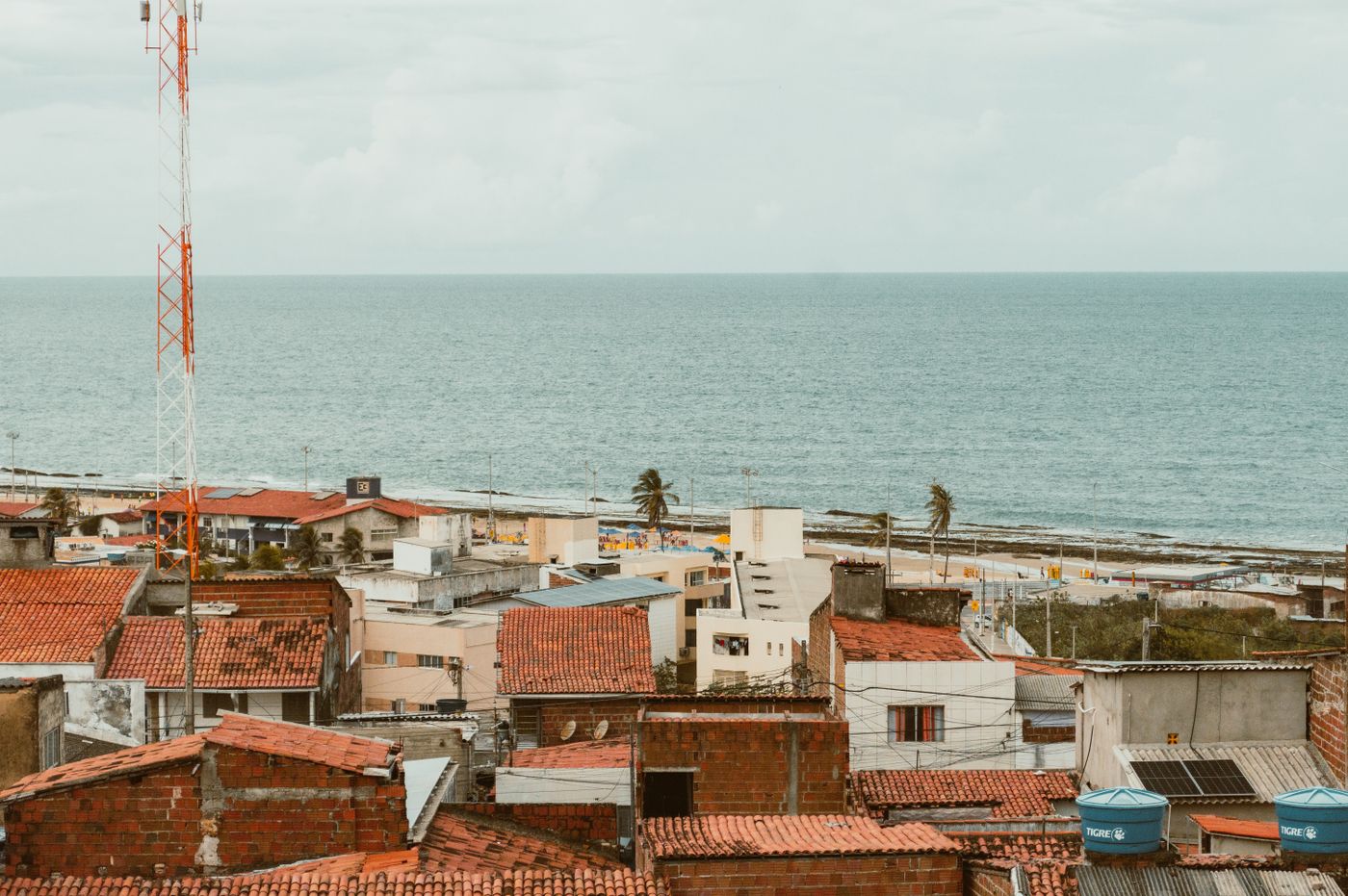 Vista aérea de telhados de cerâmica na Zona Norte de Natal, com o mar ao fundo e uma torre de antena em primeiro plano