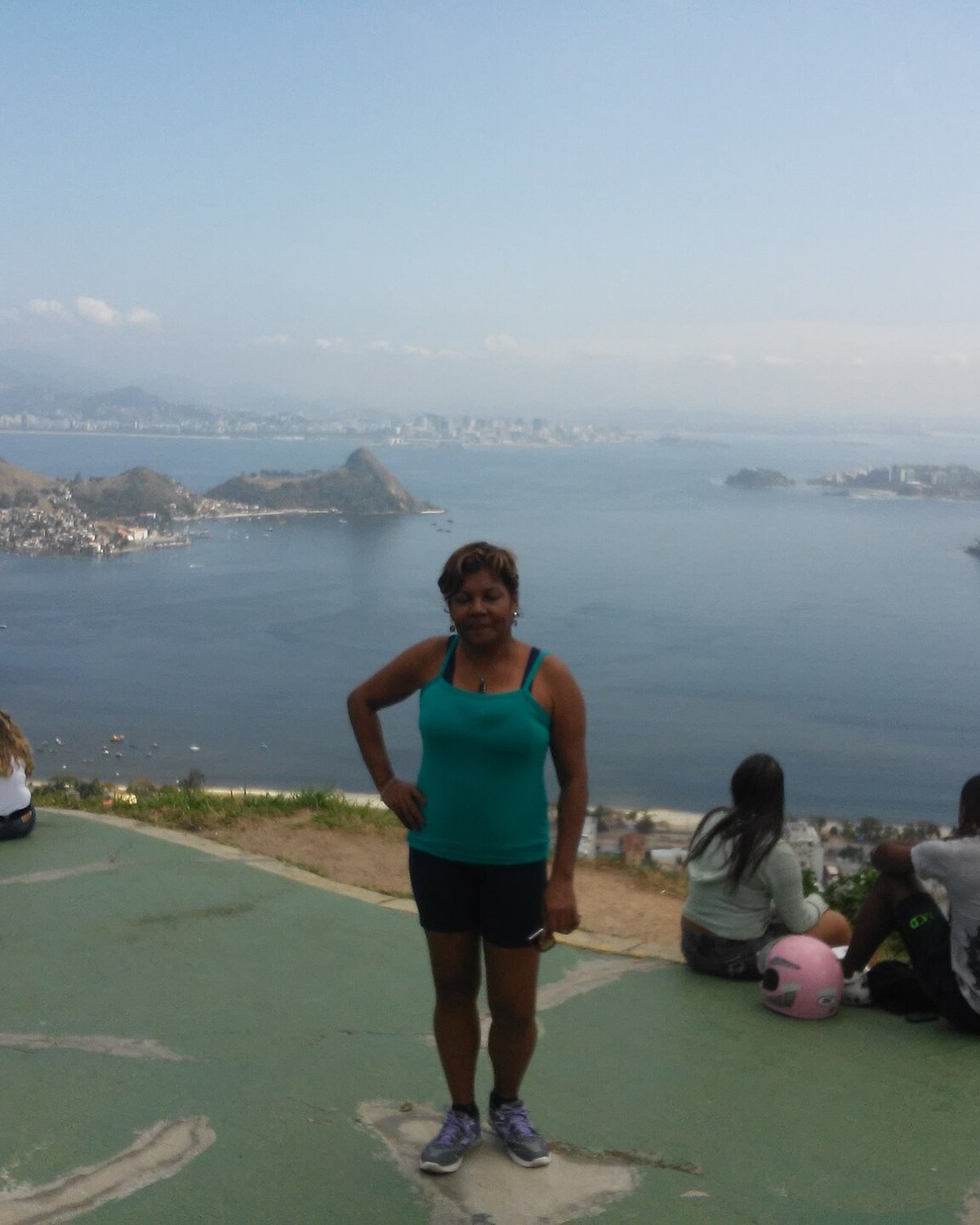 Mulher de regata verde e bermuda preta posando na rampa de voo livre do Parque da Cidade, em Niterói, com outras pessoas sentadas no chão, a Baía de Guanabara e o Rio de Janeiro ao fundo.
