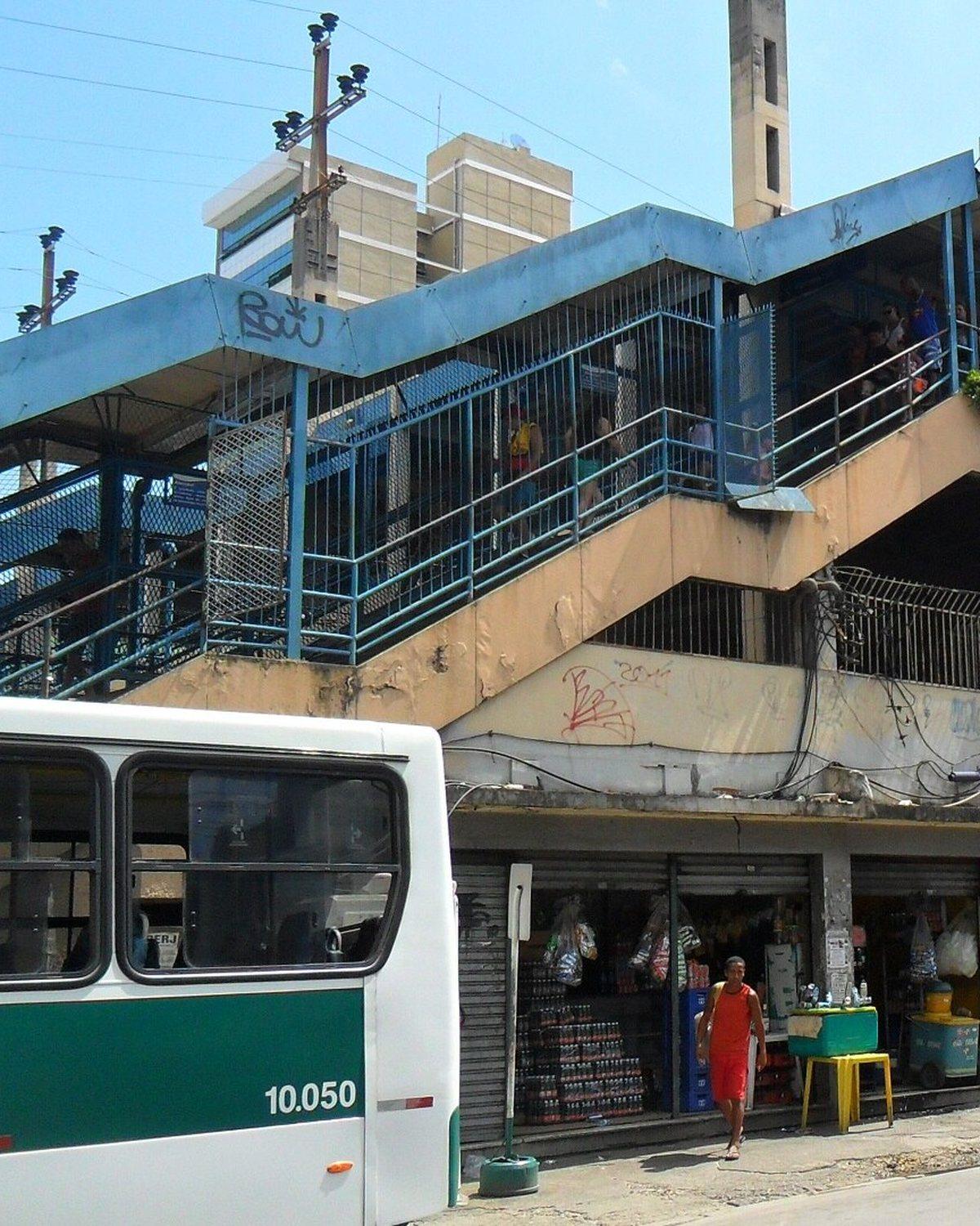 Estação ferroviária de Nova Iguaçu vista da rua, com a passarela azul, lojas no térreo, pessoas na calçada e ônibus parados no ponto em frente.