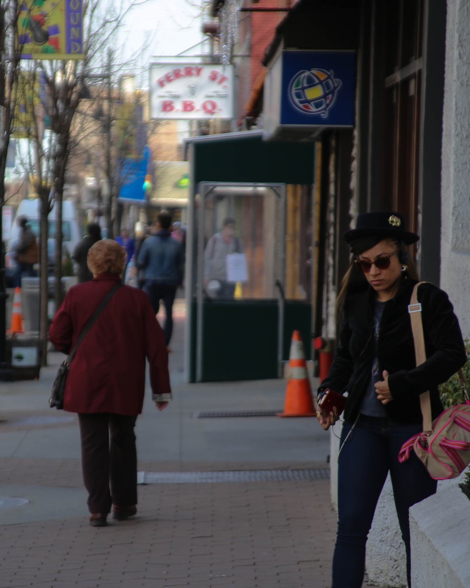 Duas mulheres caminhando pela calçada da Ferry Street, no Ironbound, em Newark, entre lojas, placas de restaurante e um cone laranja na beira da rua.
