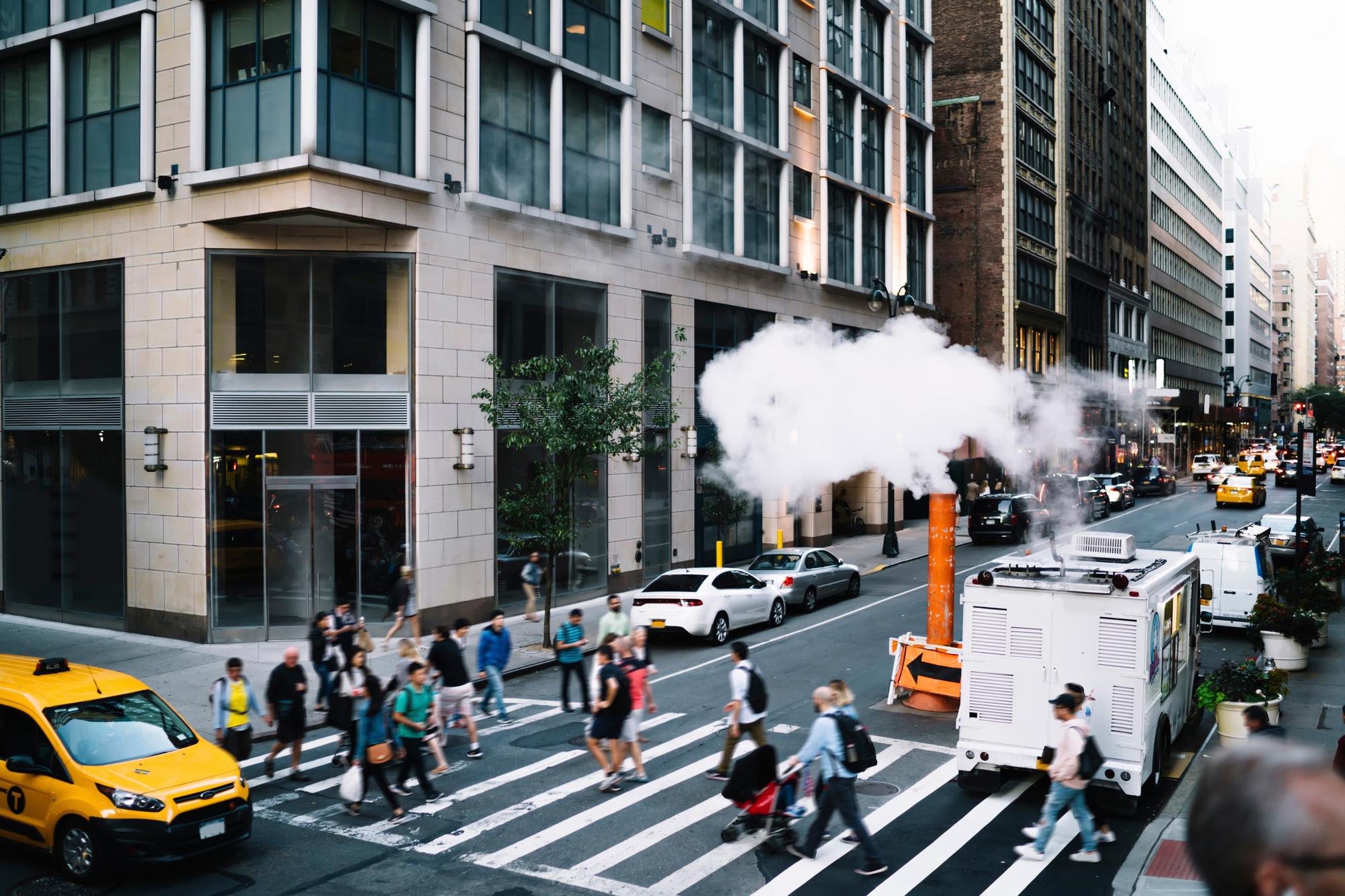 Pedestres atravessando a faixa numa esquina de Midtown, em Manhattan, com um táxi amarelo e vapor saindo da chaminé laranja no meio da rua.