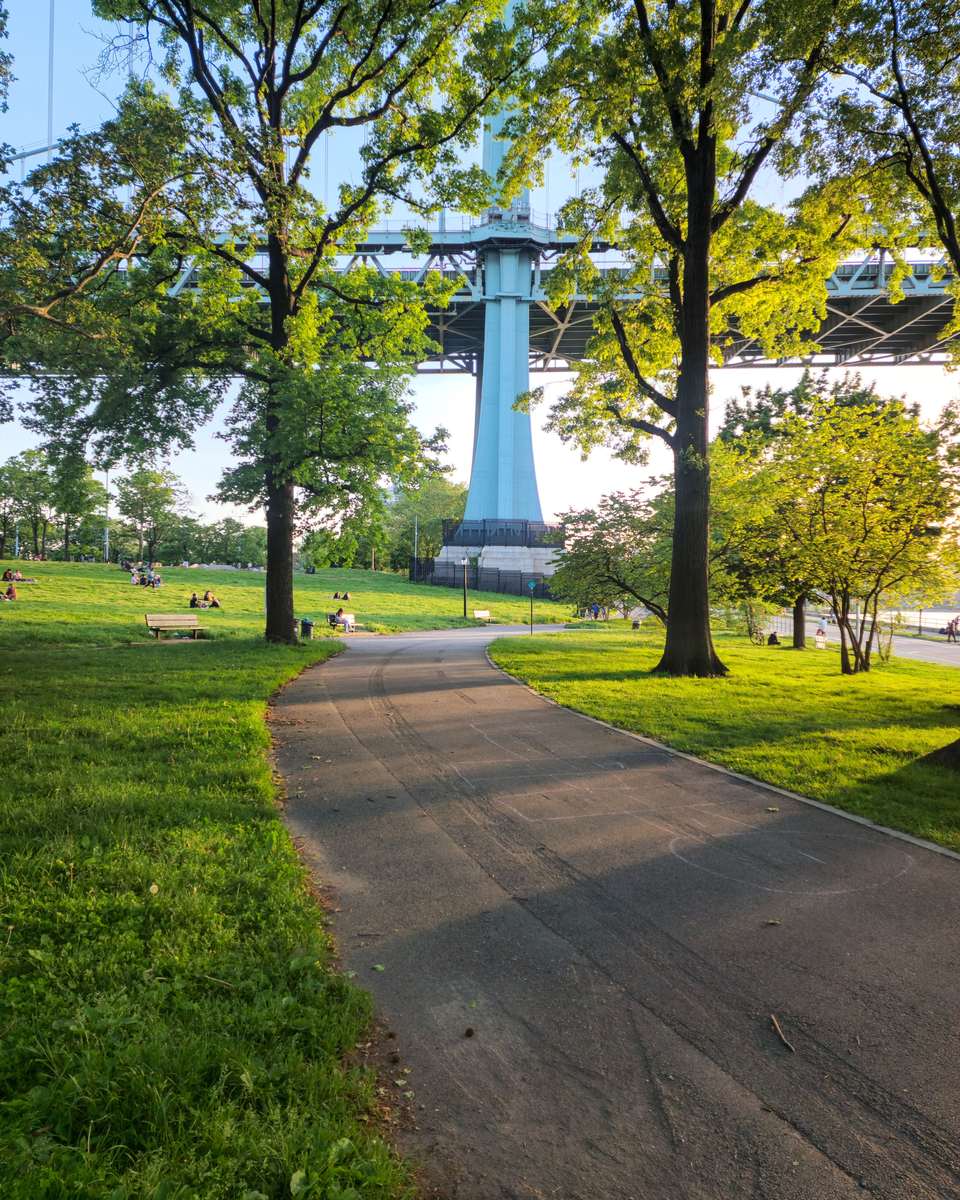 Caminho entre árvores no Astoria Park, no Queens, com gente sentada na grama e o pilar azul da ponte RFK ao fundo.