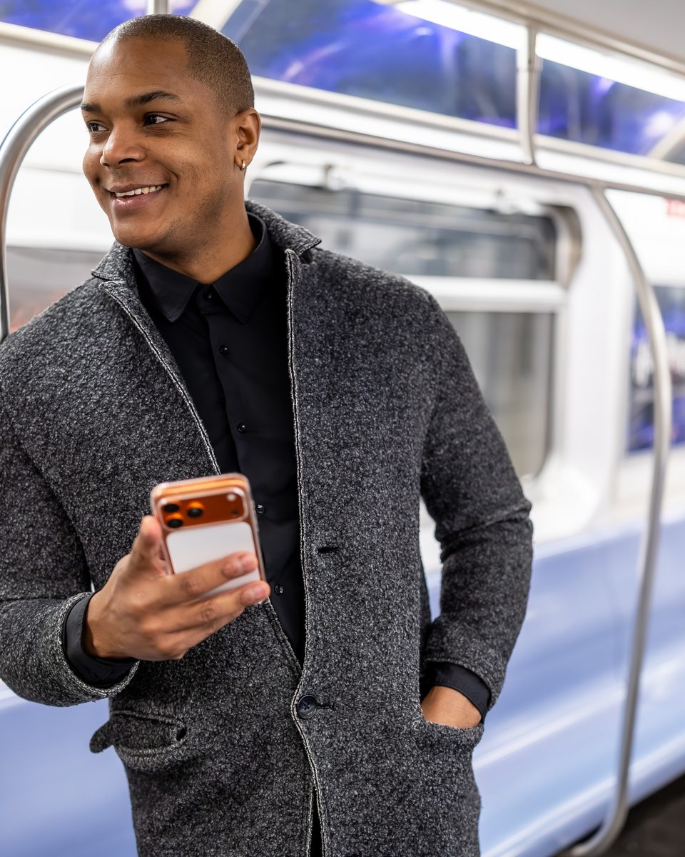 Homem sorrindo com o celular na mão dentro de um vagão do metrô de Nova York, com os bancos azuis ao fundo.